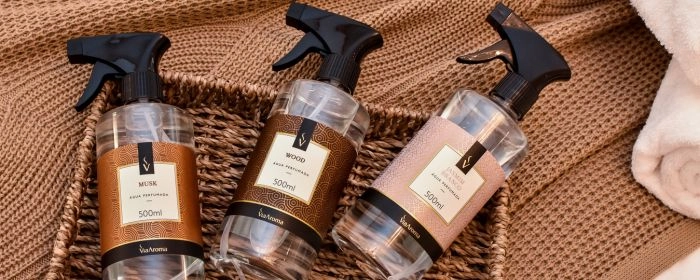 Três frascos de 500 ML de Água perfumada em um pequeno cesto marrom. Embaixo do cesto, um pano marrom com detalhes tricô
