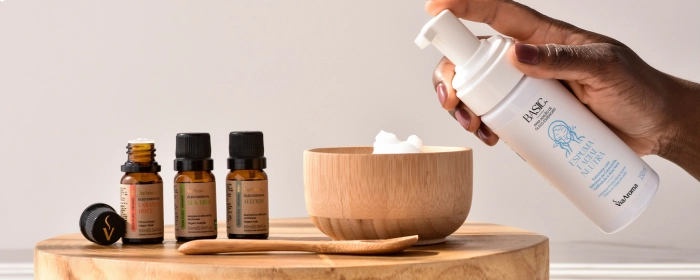 Mão despejando Espuma Facial Neutra em um pote amadeirado. Ao lado, três pequenos frescos de óleo Via Aroma.