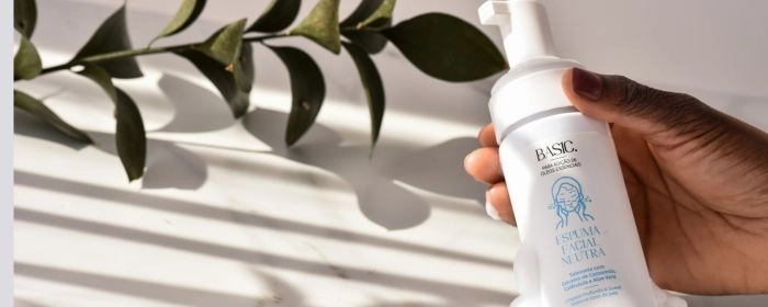 Mão de uma pessoa segurando um frasco de espuma facial Via Aroma. Plantas ao fundo.