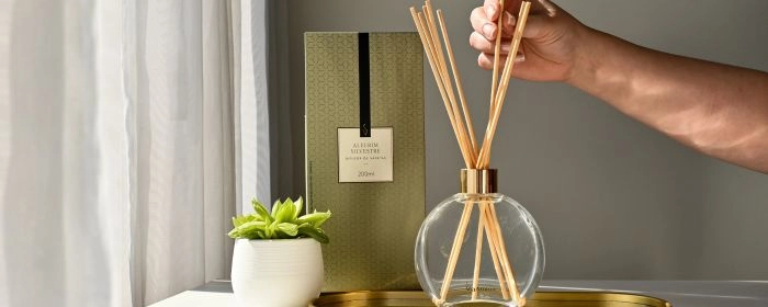 Mão realizando ajustes no Difusor de Varetas Via Aroma. Ao fundo, uma caixa de um produto da Via Aroma, com um pequeno jarro de plantas e uma cortina branca